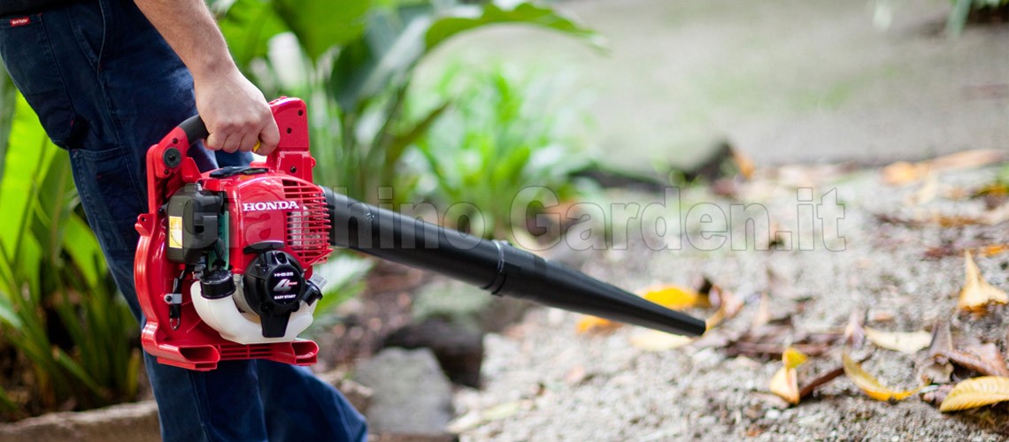 Operatore che impugna e solleva un soffiatore a scoppio rosso Honda HHB25 in un giardino, con il tubo di soffiaggio puntato verso foglie secche a terra.