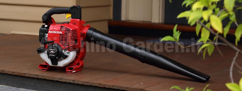 Soffiatore a scoppio rosso Honda HHB25 appoggiato su una pavimentazione esterna in legno scuro, con il lungo tubo di soffiaggio rivolto verso destra e foglie verdi in primo piano sfocate.