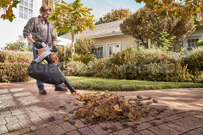 Operatore in piedi su un vialetto in mattoni mentre utilizza l'aspirafoglie STIHL SHA 56 per aspirare un cumulo di foglie secche autunnali. Sullo sfondo si vedono una casa e un giardino curato.