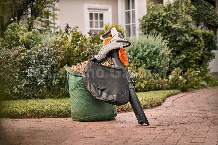 Lo STIHL SHA 56 in modalità aspirafoglie è posizionato in verticale sul vialetto di un cortile, appoggiato a un grande sacco da giardino verde riempito di fogliame sminuzzato.