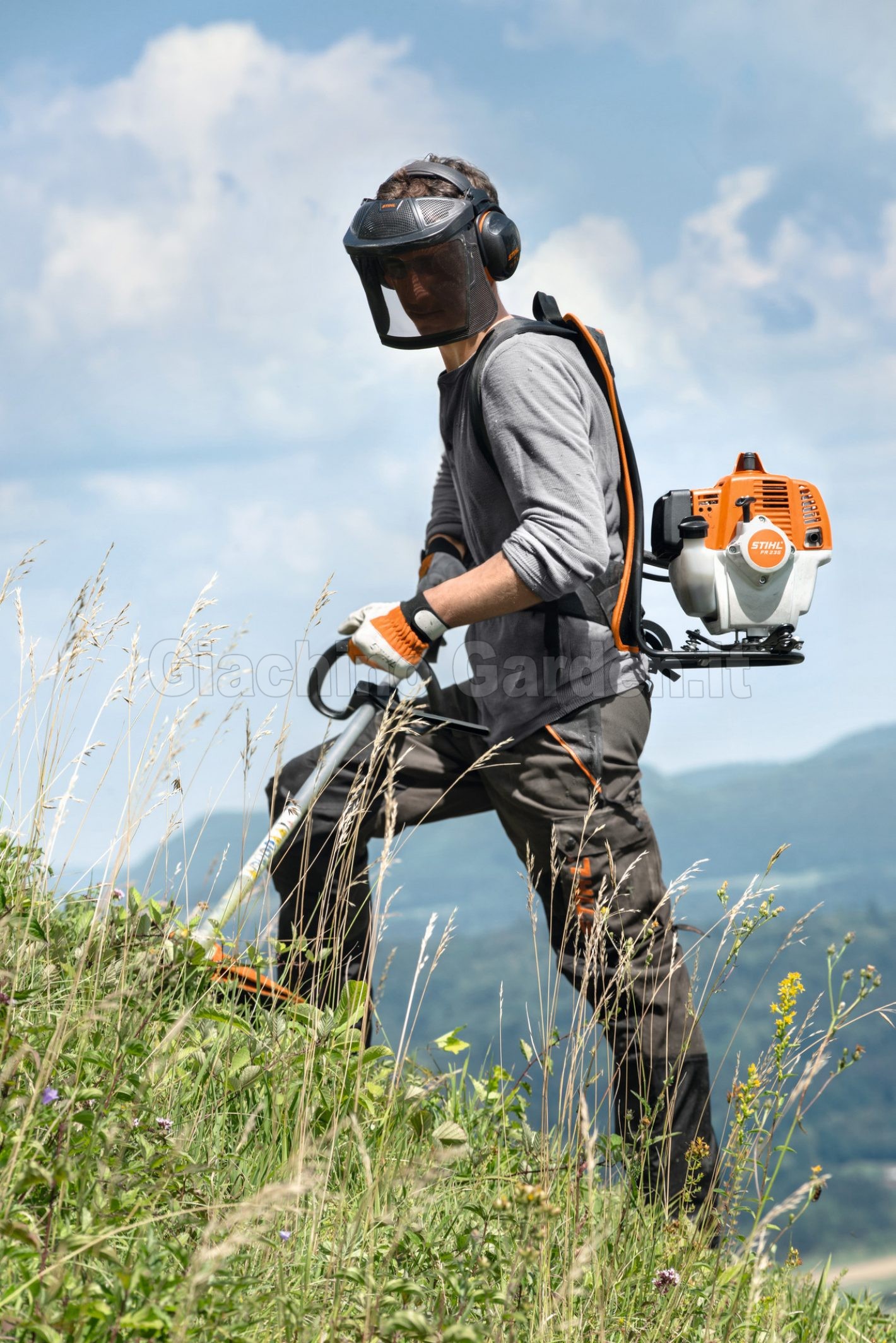 Operatore con decespugliatore spalleggiato STIHL FR 235 al lavoro su un prato in forte pendenza, mentre indossa visiera protettiva, cuffie antirumore e abbigliamento da lavoro arancione e grigio.