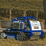 Robot sfalciatutto radiocomandato Barbieri X ROT 70 EVO blu e bianco, al lavoro su un prato con un vigneto sullo sfondo.