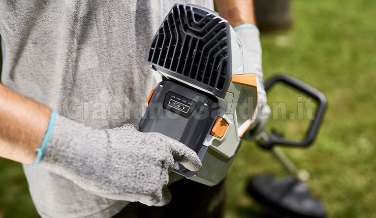 Primo piano della batteria on-board Pellenc ULiB 200E agganciata alla parte posteriore di un utensile della gamma Essential. Un operatore con guanti da lavoro grigi indica con il dito l'indicatore di carica a LED sul guscio nero della batteria compatta. Sullo sfondo sfocato si vede un prato verde