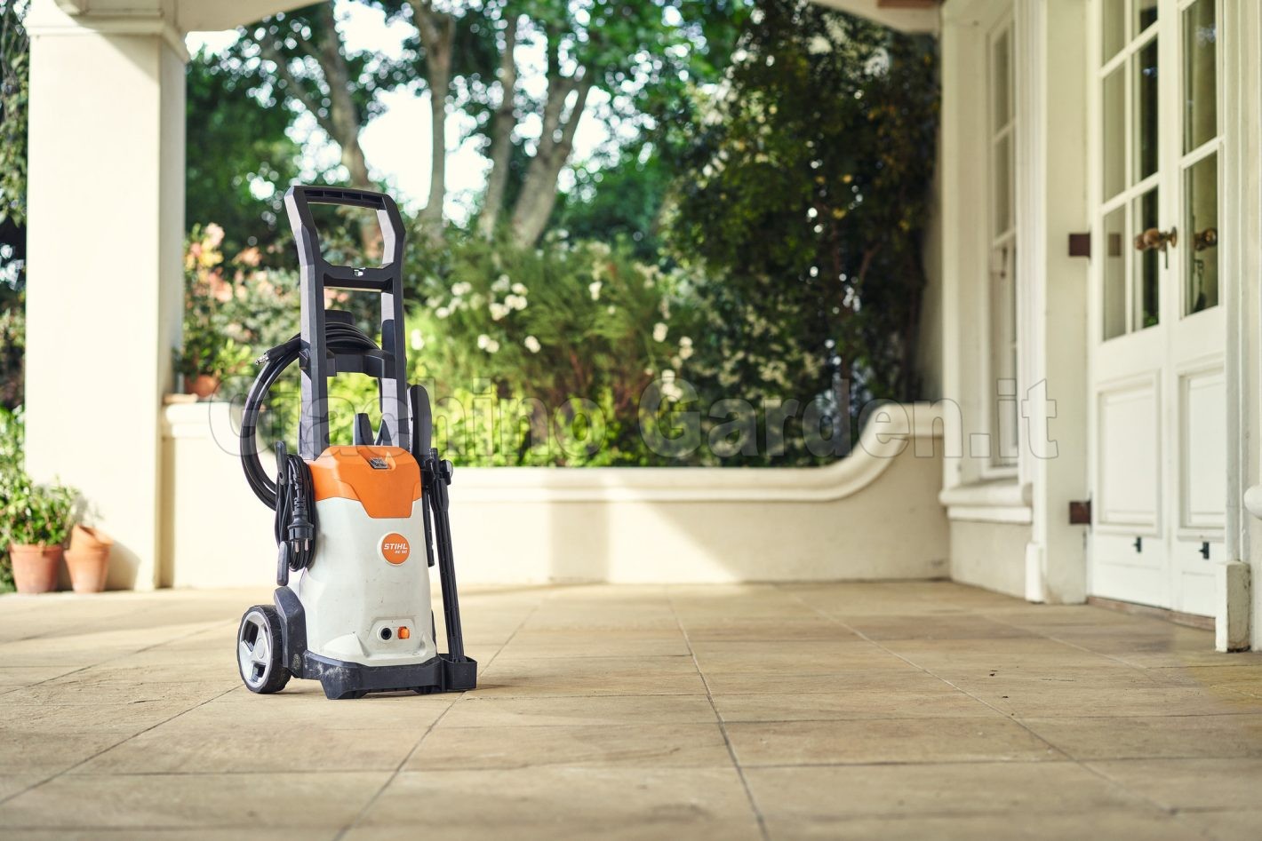 Idropulitrice compatta STIHL RE 90 posizionata sul pavimento piastrellato di un portico domestico esterno, con piante, alberi e una porta a vetri bianca sullo sfondo.