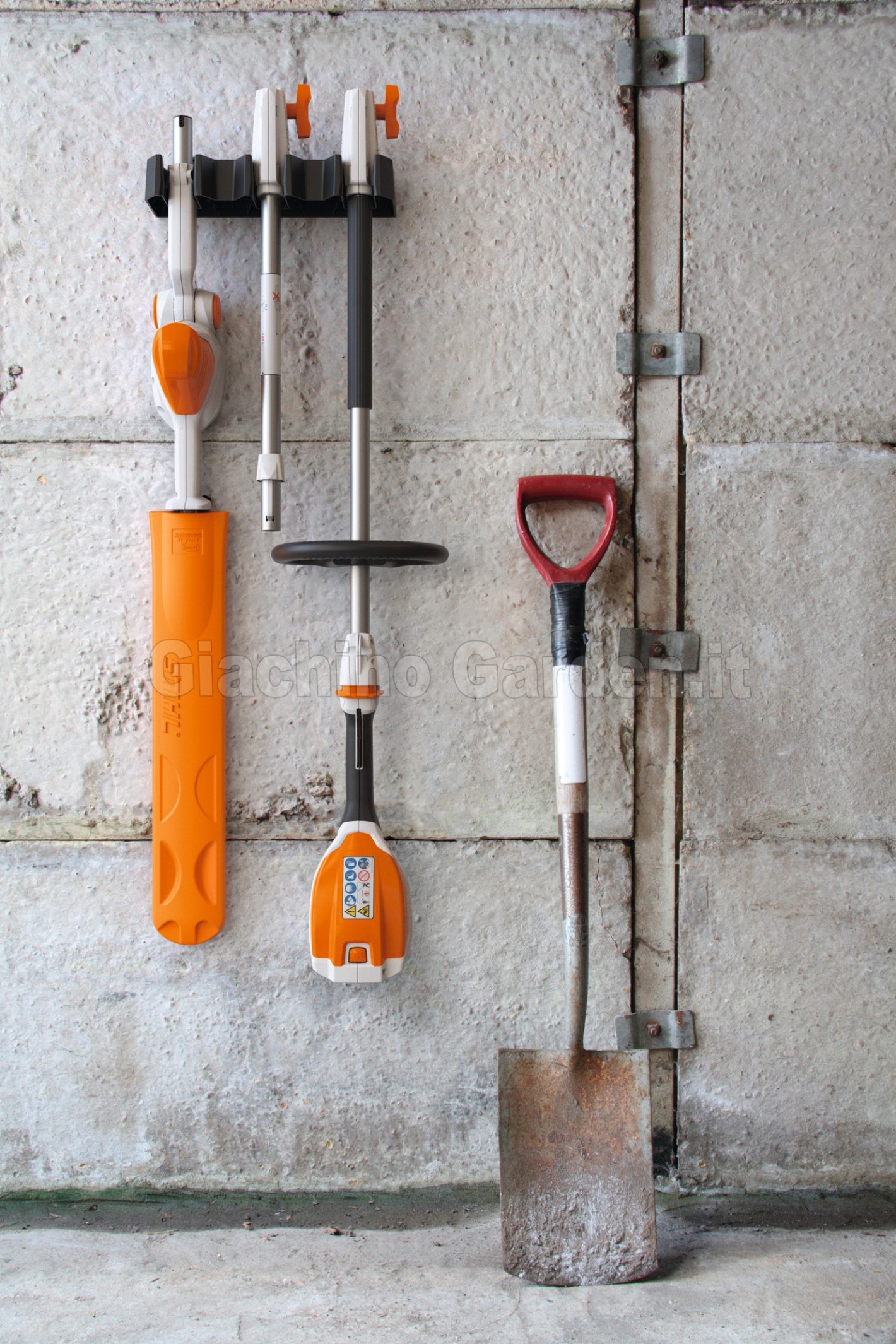 Componenti del tagliasiepi telescopico a batteria STIHL HLA 56 smontati e appesi a un supporto a muro in garage, accanto a una vanga da giardino.